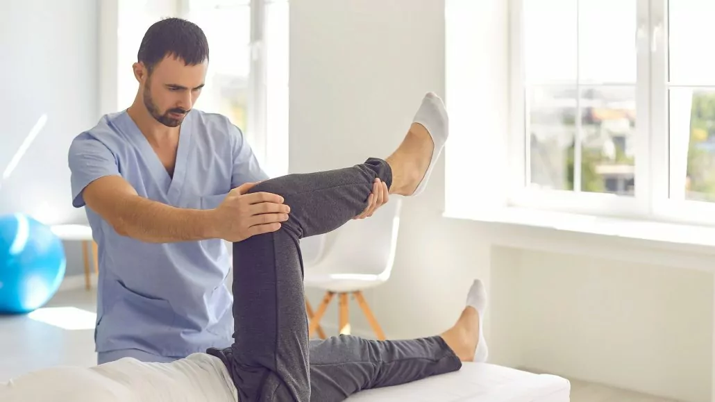 Therapeut macht mit dem Patient Knie-Rehaübungen nach einer Knie-OP auf der Liege.